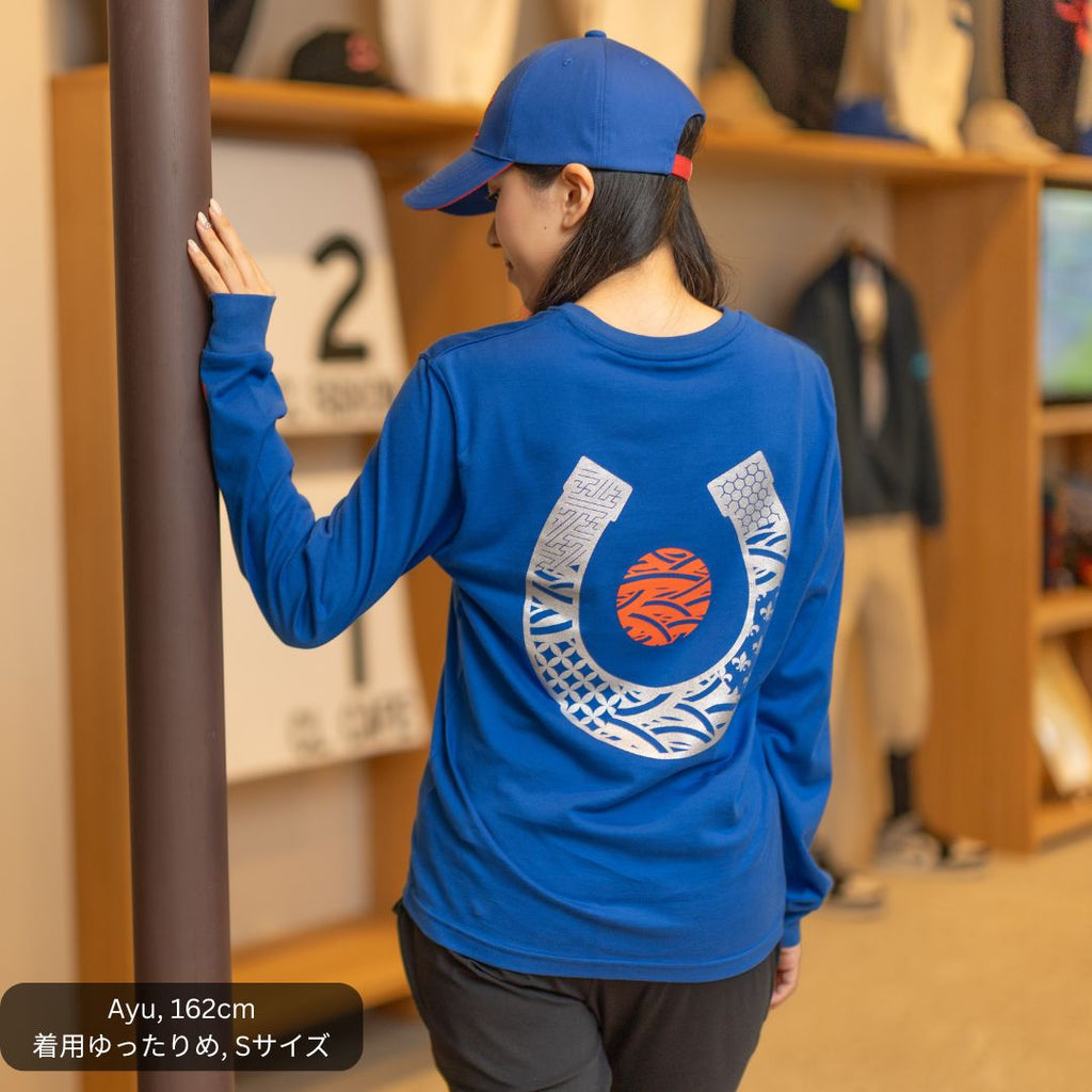 女性モデルがCL by C.ルメールの「LONG TETSU」ブルーを着用。背面には和柄で構成された大きなシルバーの蹄鉄モチーフと、日の丸をイメージした赤い円形デザインを配置。日本製ロングスリーブTシャツとブルーのキャップを合わせた、競馬と日本文化を融合したスタイリング。