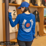 女性モデルがCL by C.ルメールの「LONG TETSU」ブルーを着用。背面には和柄で構成された大きなシルバーの蹄鉄モチーフと、日の丸をイメージした赤い円形デザインを配置。日本製ロングスリーブTシャツとブルーのキャップを合わせた、競馬と日本文化を融合したスタイリング。