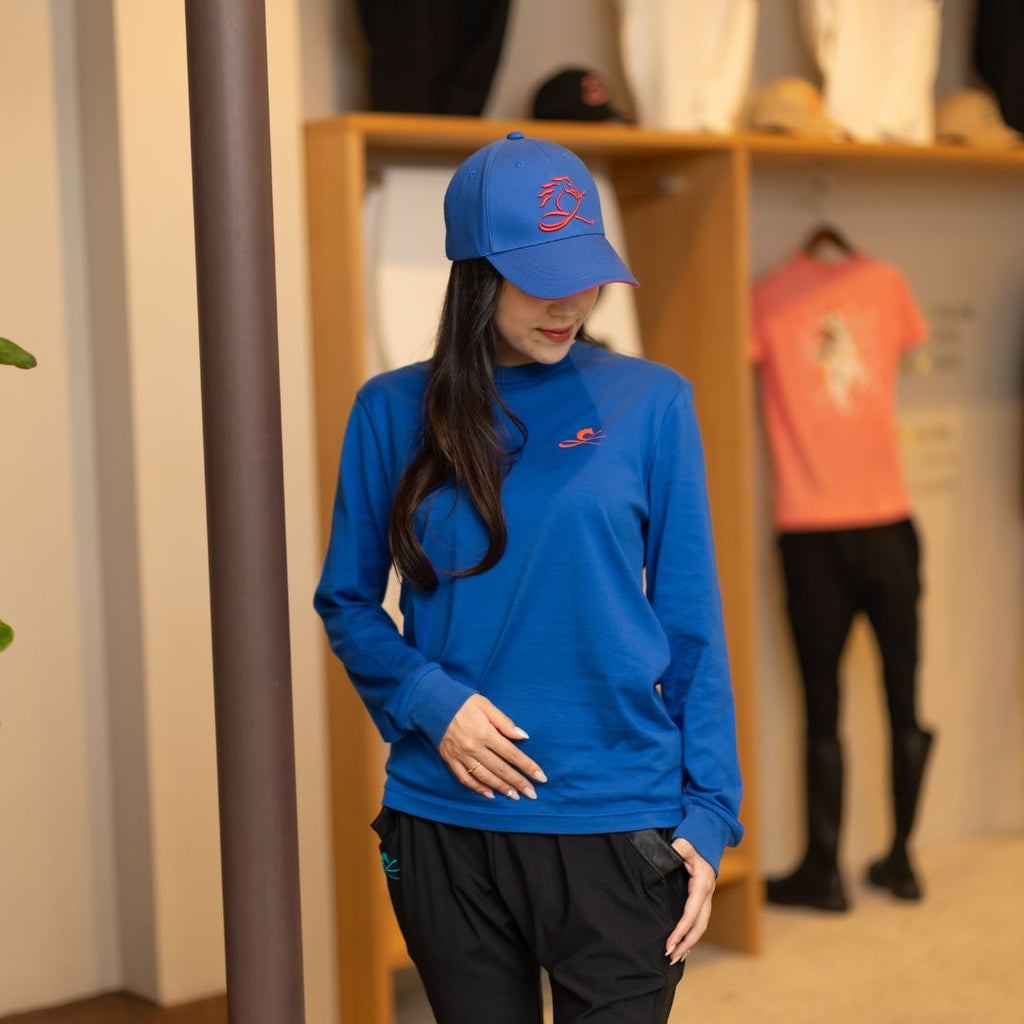 女性モデルがCL by C.ルメールの「RED HORSE CAP」を着用。鮮やかなブルーのキャップのフロントには赤い馬の刺繍ロゴを配置し、つば裏とアジャスターにも赤のアクセントを採用。日本製のロングスリーブTシャツと合わせた、競馬をテーマにしたスポーティーなスタイリング。